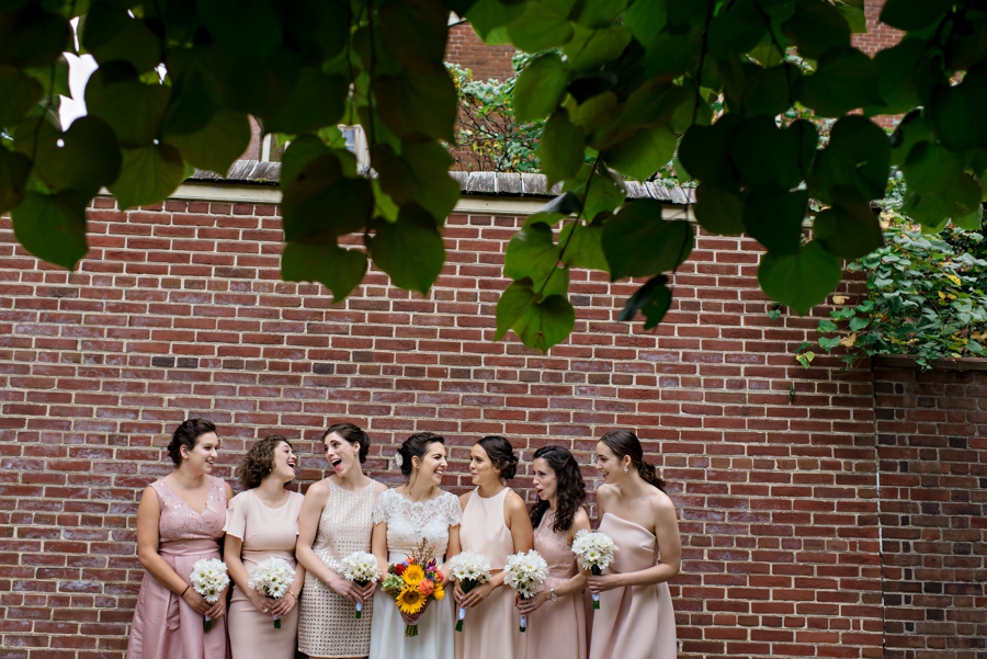 A bride laughing with her bridesmaids agains a brick wall.